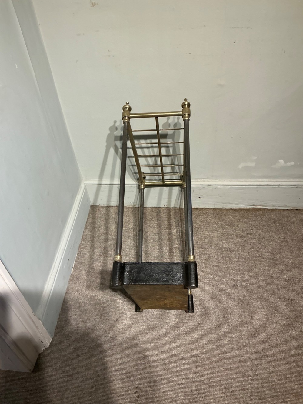 An antique brass and steel stick stand featuring a six-section top grid, urn finials, tubular uprights, and a black cast iron drip tray base. View VI
