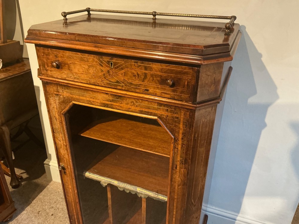 A Victorian burr walnut music cabinet featuring inlaid boxwood stringing, a top drawer, a glazed door revealing internal shelves and vertical dividers, and a brass gallery rail. View V