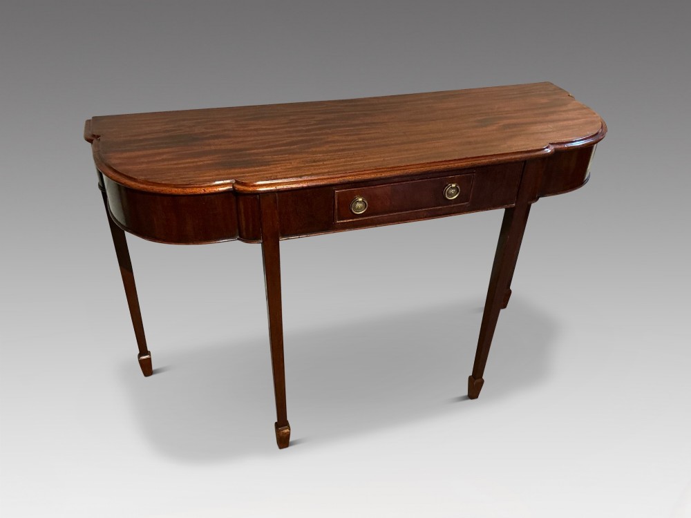 An antique mahogany hall table featuring a serpentine front, a single central drawer with brass ring handles, and four tapered square legs ending in spade feet.