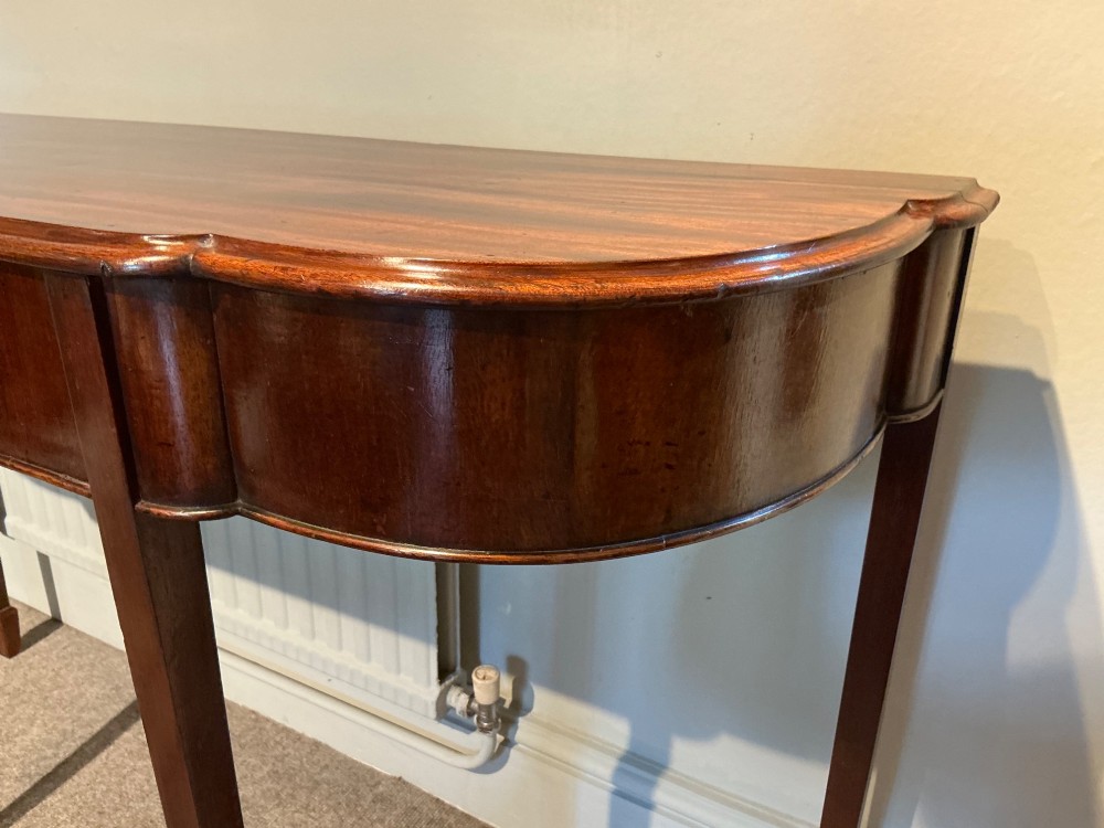 An antique mahogany hall table featuring a serpentine front, a single central drawer with brass ring handles, and four tapered square legs ending in spade feet. View IV