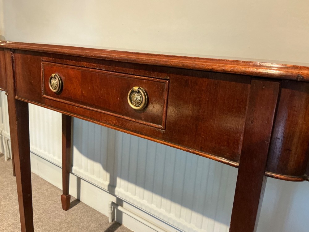 An antique mahogany hall table featuring a serpentine front, a single central drawer with brass ring handles, and four tapered square legs ending in spade feet. View VI