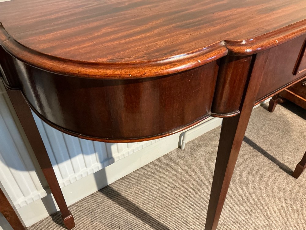 An antique mahogany hall table featuring a serpentine front, a single central drawer with brass ring handles, and four tapered square legs ending in spade feet. View VIII