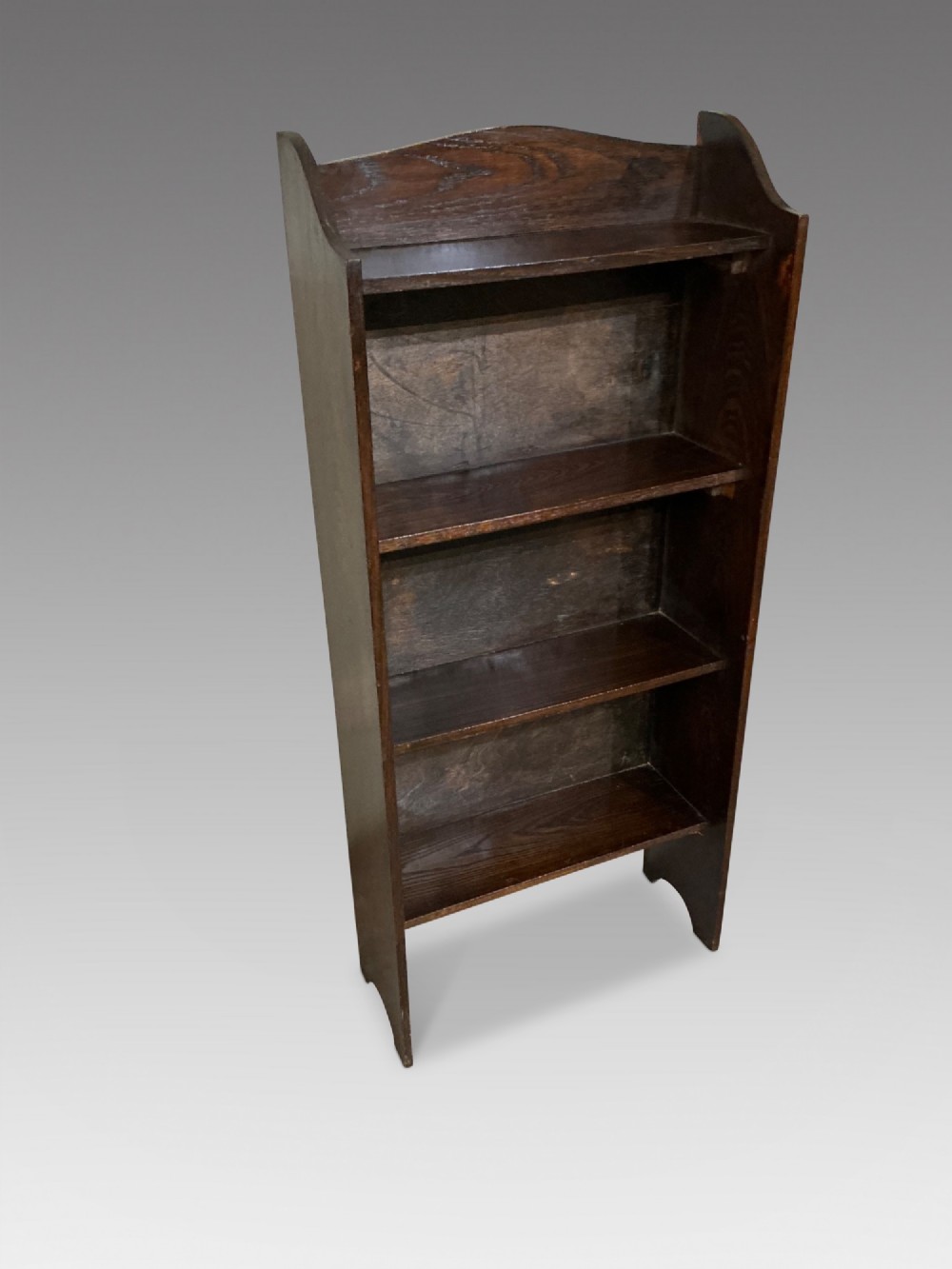 An antique dark oak open bookcase featuring four fixed shelves, cutout side panels forming feet, and a curved top cresting rail.