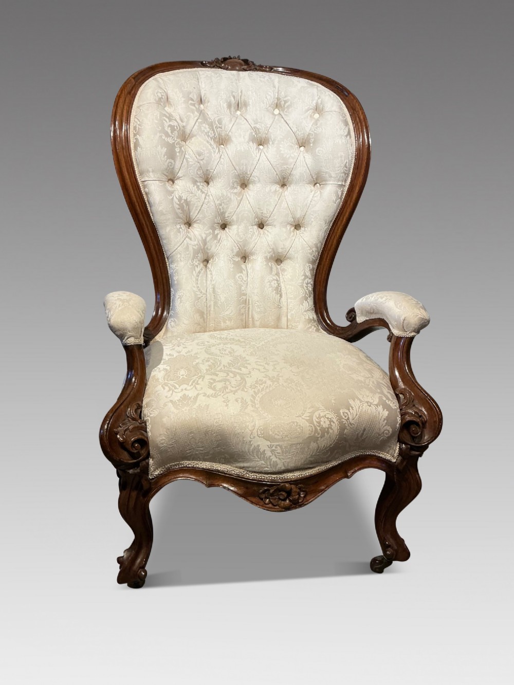 A Victorian high-back armchair featuring a carved dark wood frame, deep button-tufted cream damask upholstery, and cabriole legs on brass castors.