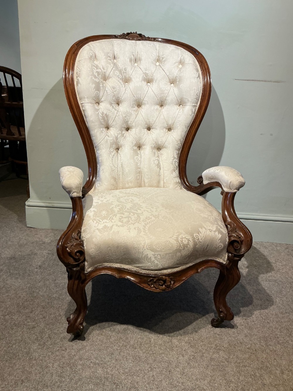A Victorian high-back armchair featuring a carved dark wood frame, deep button-tufted cream damask upholstery, and cabriole legs on brass castors. View II