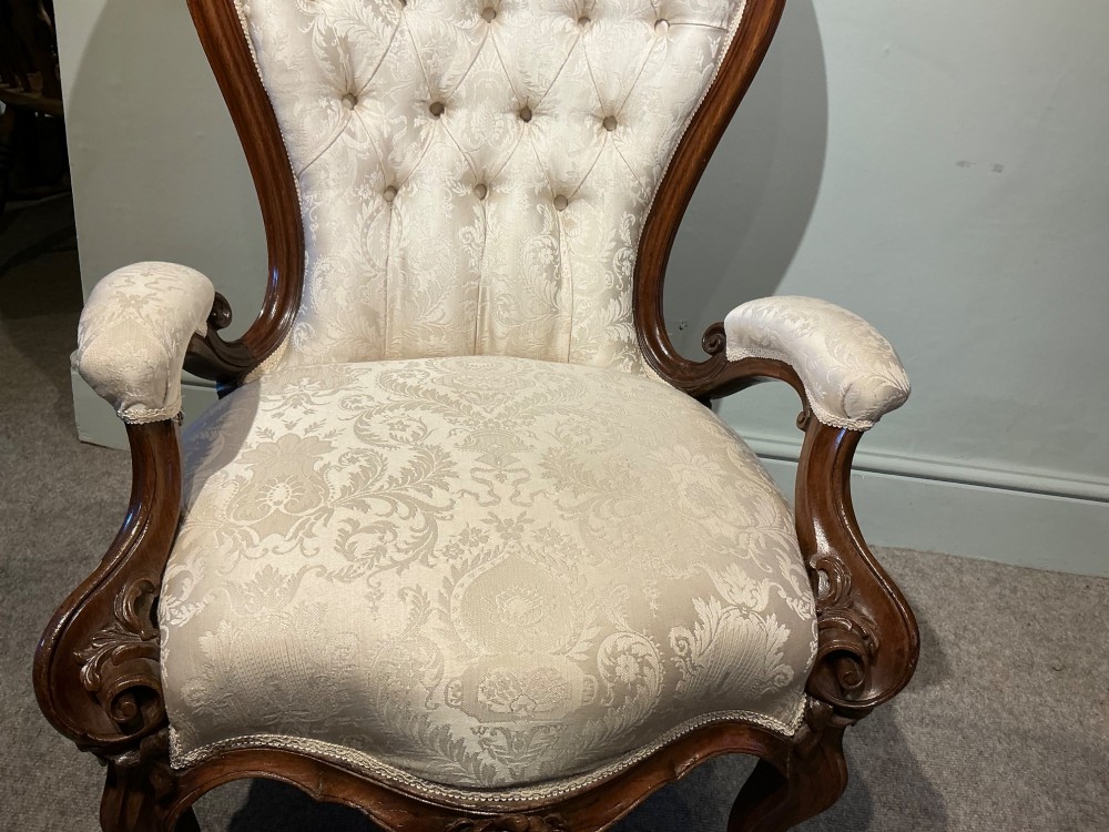 A Victorian high-back armchair featuring a carved dark wood frame, deep button-tufted cream damask upholstery, and cabriole legs on brass castors. View IV