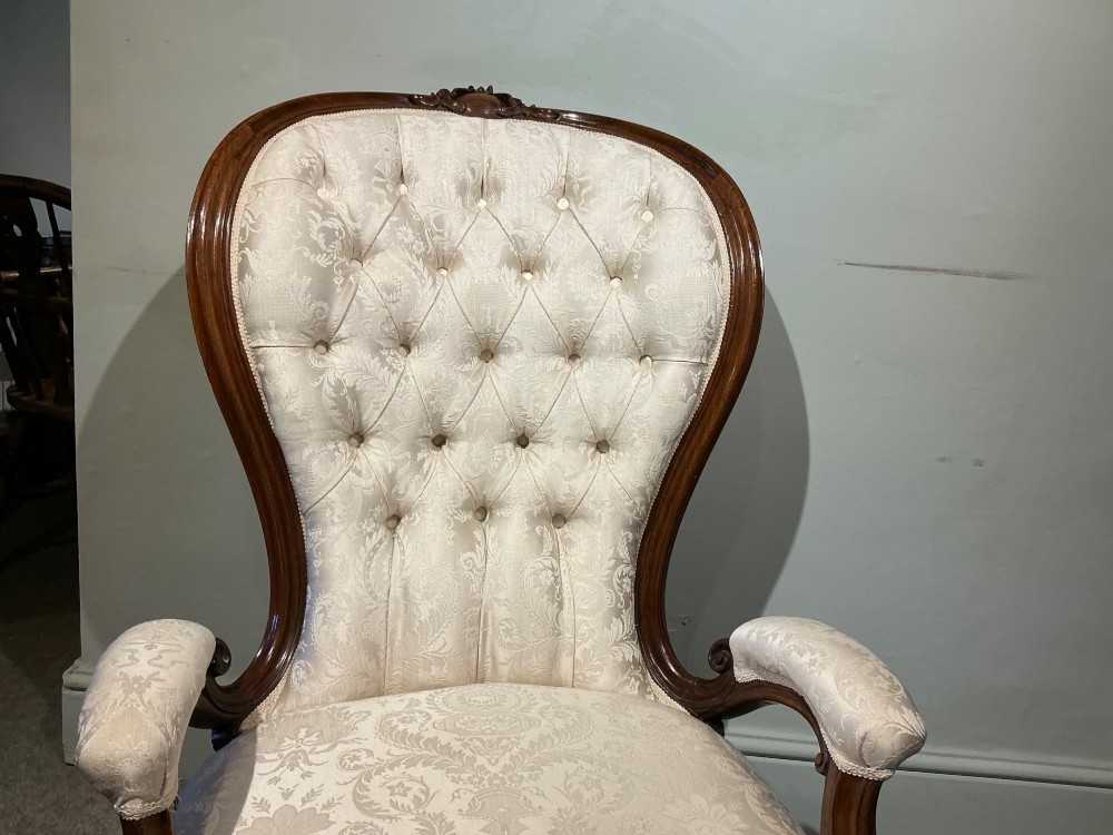 A Victorian high-back armchair featuring a carved dark wood frame, deep button-tufted cream damask upholstery, and cabriole legs on brass castors. View V
