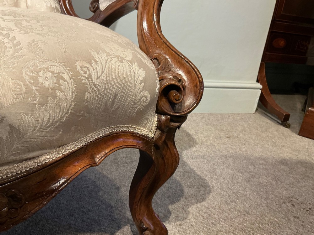 A Victorian high-back armchair featuring a carved dark wood frame, deep button-tufted cream damask upholstery, and cabriole legs on brass castors. View VI