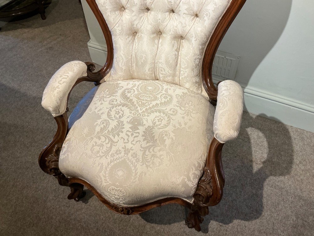 A Victorian high-back armchair featuring a carved dark wood frame, deep button-tufted cream damask upholstery, and cabriole legs on brass castors. View IX