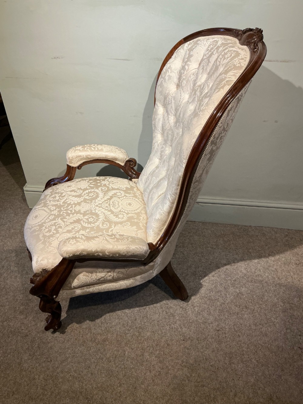 A Victorian high-back armchair featuring a carved dark wood frame, deep button-tufted cream damask upholstery, and cabriole legs on brass castors. View XI