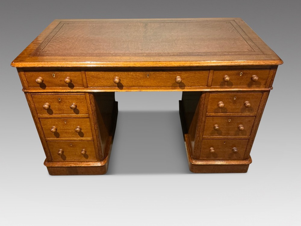 An antique oak pedestal desk featuring a rectangular leather inset top, nine drawers with turned wooden knobs, and twin plinth bases.