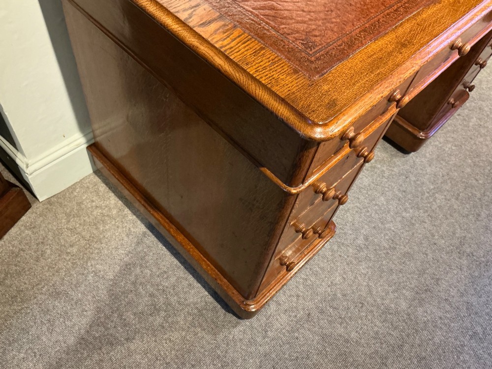An antique oak pedestal desk featuring a rectangular leather inset top, nine drawers with turned wooden knobs, and twin plinth bases. View IV