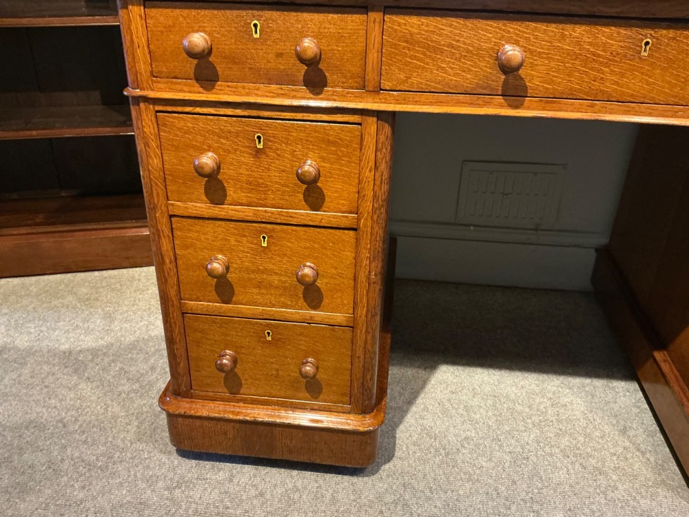 An antique oak pedestal desk featuring a rectangular leather inset top, nine drawers with turned wooden knobs, and twin plinth bases. View V