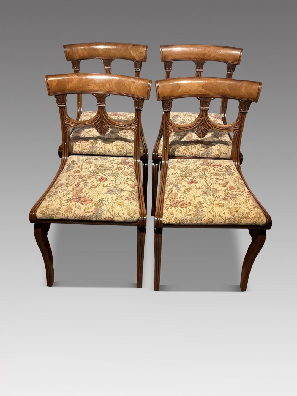 A set of four antique mahogany dining chairs with carved palmette back splats, curved crest rails, sabre legs, and floral tapestry upholstered drop-in seats.