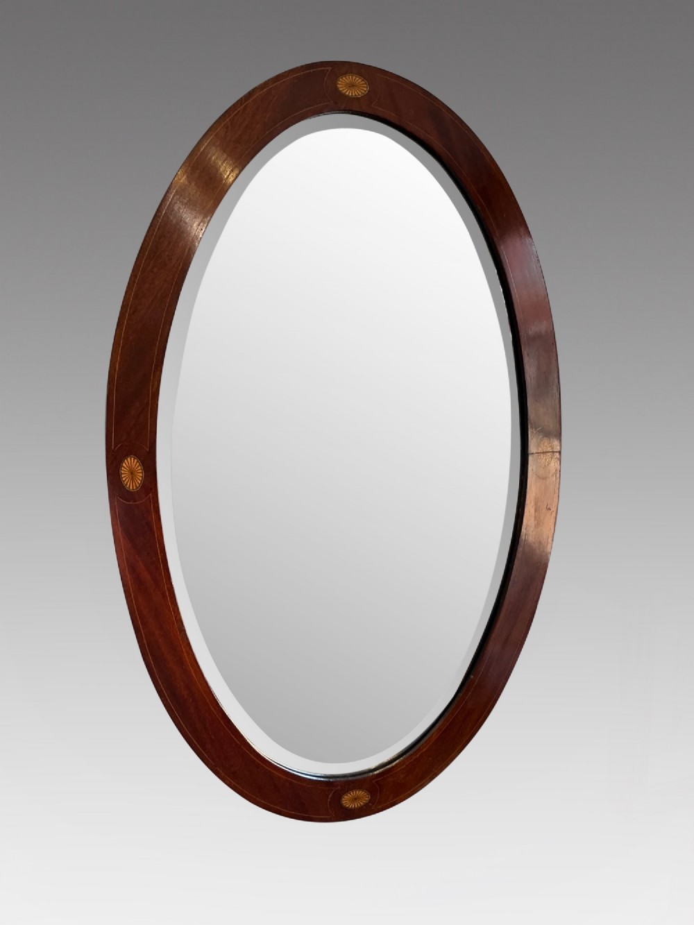 An antique oval mahogany wall mirror featuring a polished wooden frame accented with stringing inlay and four decorative marquetry medallions surrounding a bevelled mirror plate.