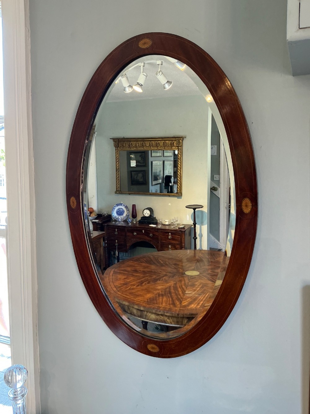 An antique oval mahogany wall mirror featuring a polished wooden frame accented with stringing inlay and four decorative marquetry medallions surrounding a bevelled mirror plate. View II