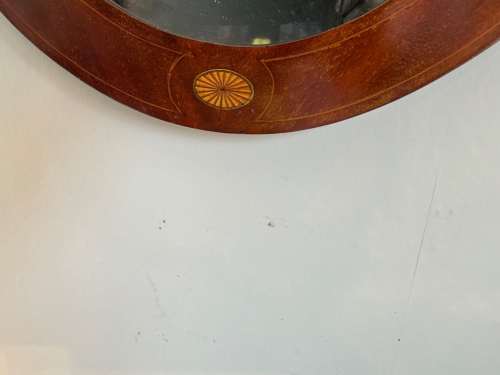 An antique oval mahogany wall mirror featuring a polished wooden frame accented with stringing inlay and four decorative marquetry medallions surrounding a bevelled mirror plate. View IV