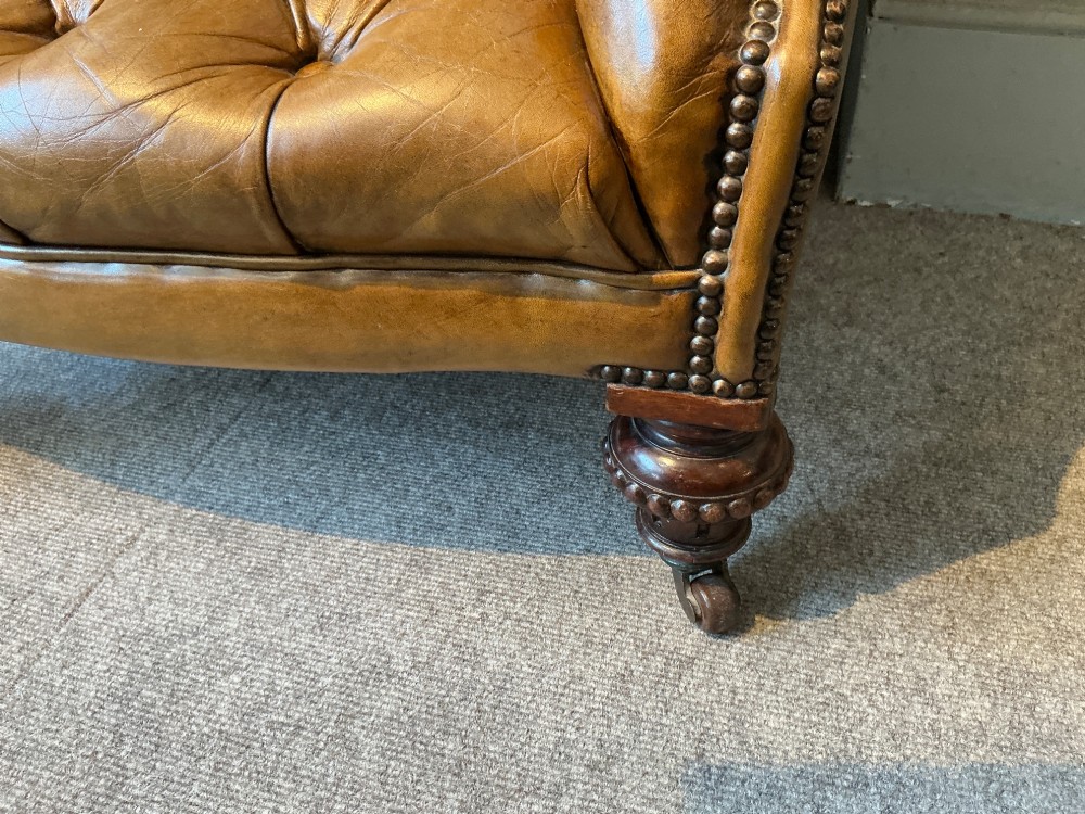 A Victorian warm brown leather armchair featuring deep button tufting, brass stud detailing, and turned wood front legs on castors. View V