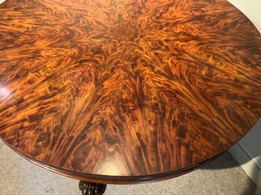 A circular antique mahogany centre table featuring a radially segmented flame veneer top supported by a central pedestal with carved claw feet. View V