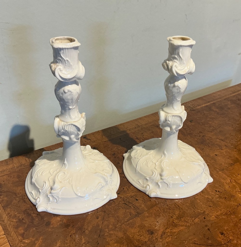 A pair of off-white glazed porcelain candlesticks featuring sculpted Rococo scrollwork and shell motifs on wide circular bases.