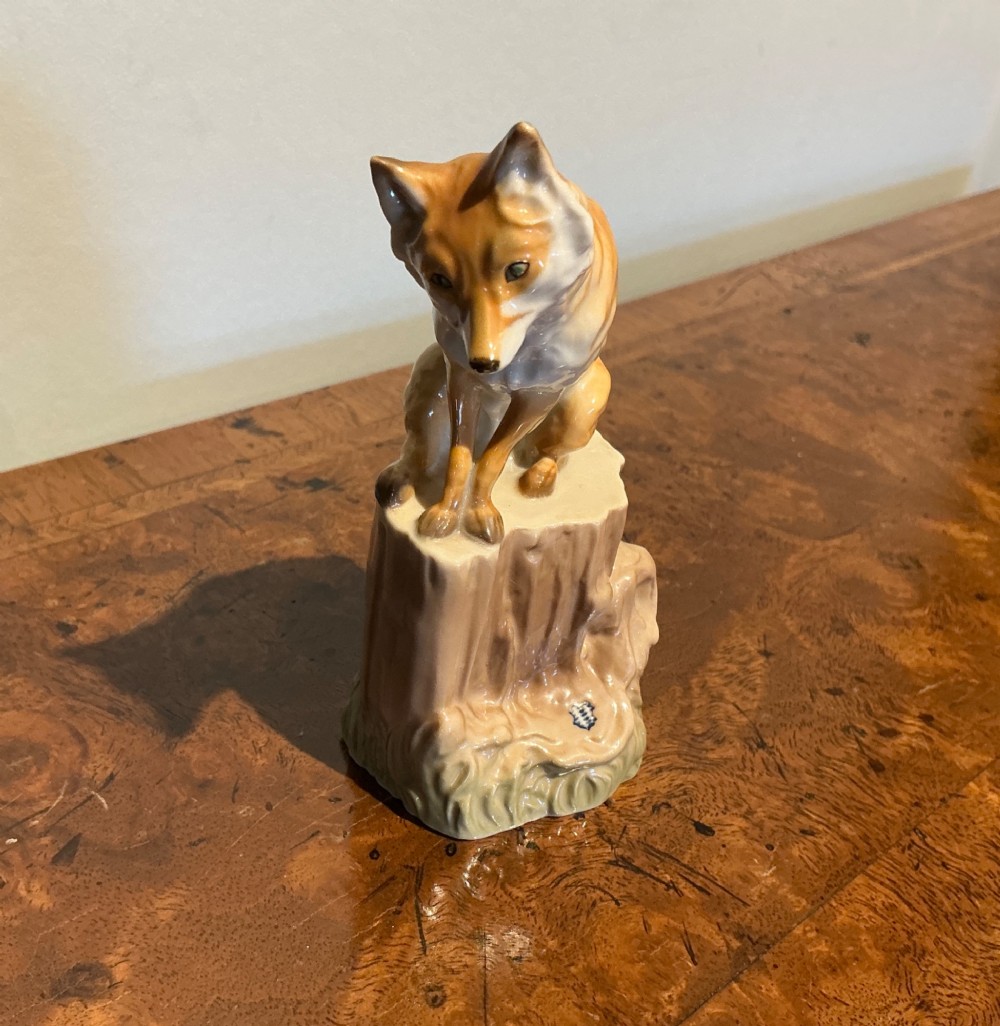 A glazed porcelain figurine depicting a rust-orange fox seated atop a moulded tree stump base with hand-painted details.