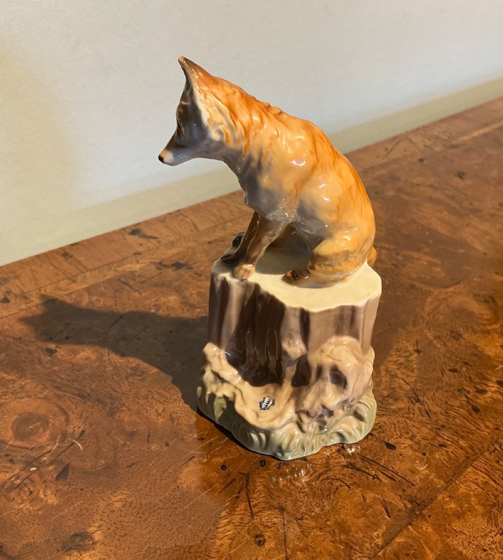A glazed porcelain figurine depicting a rust-orange fox seated atop a moulded tree stump base with hand-painted details. View II