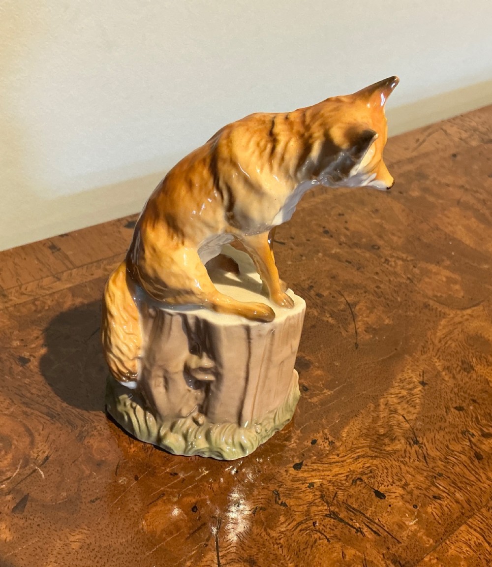A glazed porcelain figurine depicting a rust-orange fox seated atop a moulded tree stump base with hand-painted details. View IV