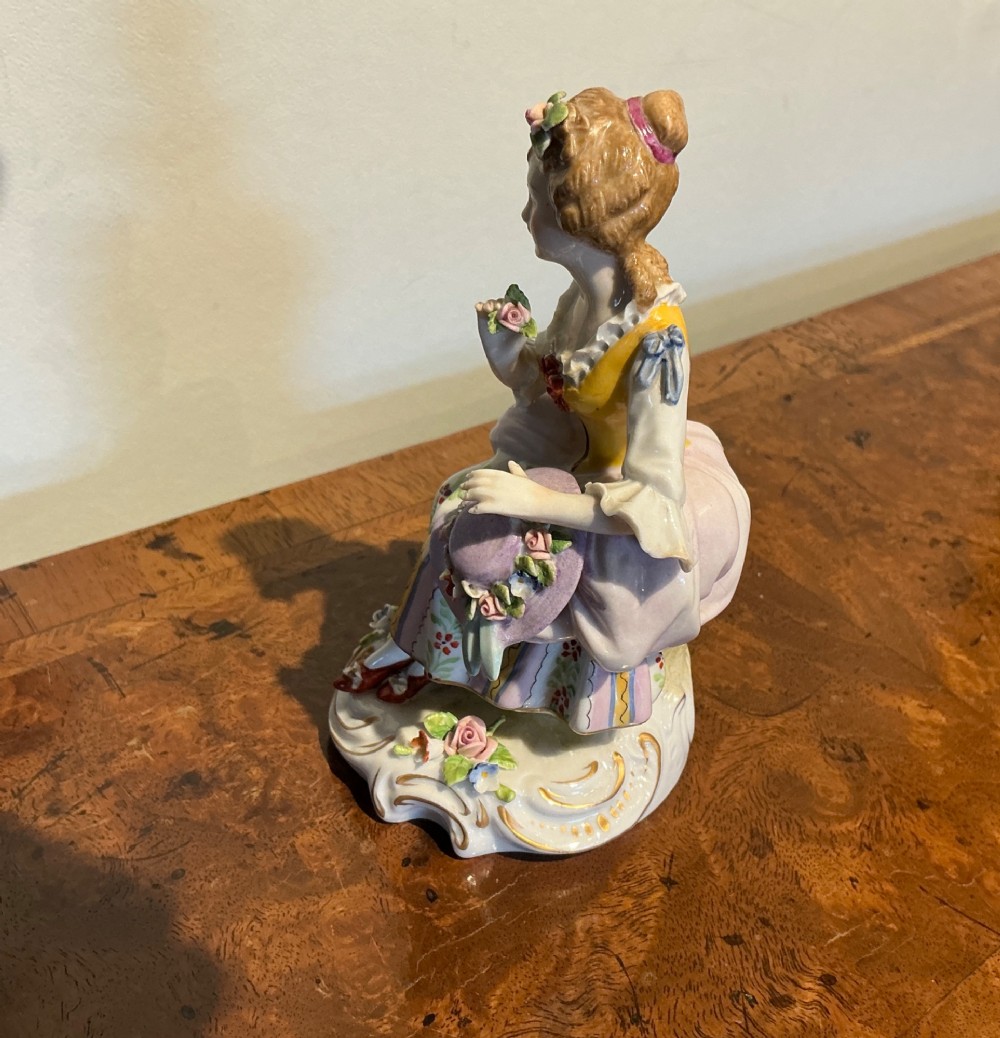 A Sitzendorf porcelain figurine depicting a seated woman in a floral skirt holding a flower-adorned bonnet on a scrolled base. View II