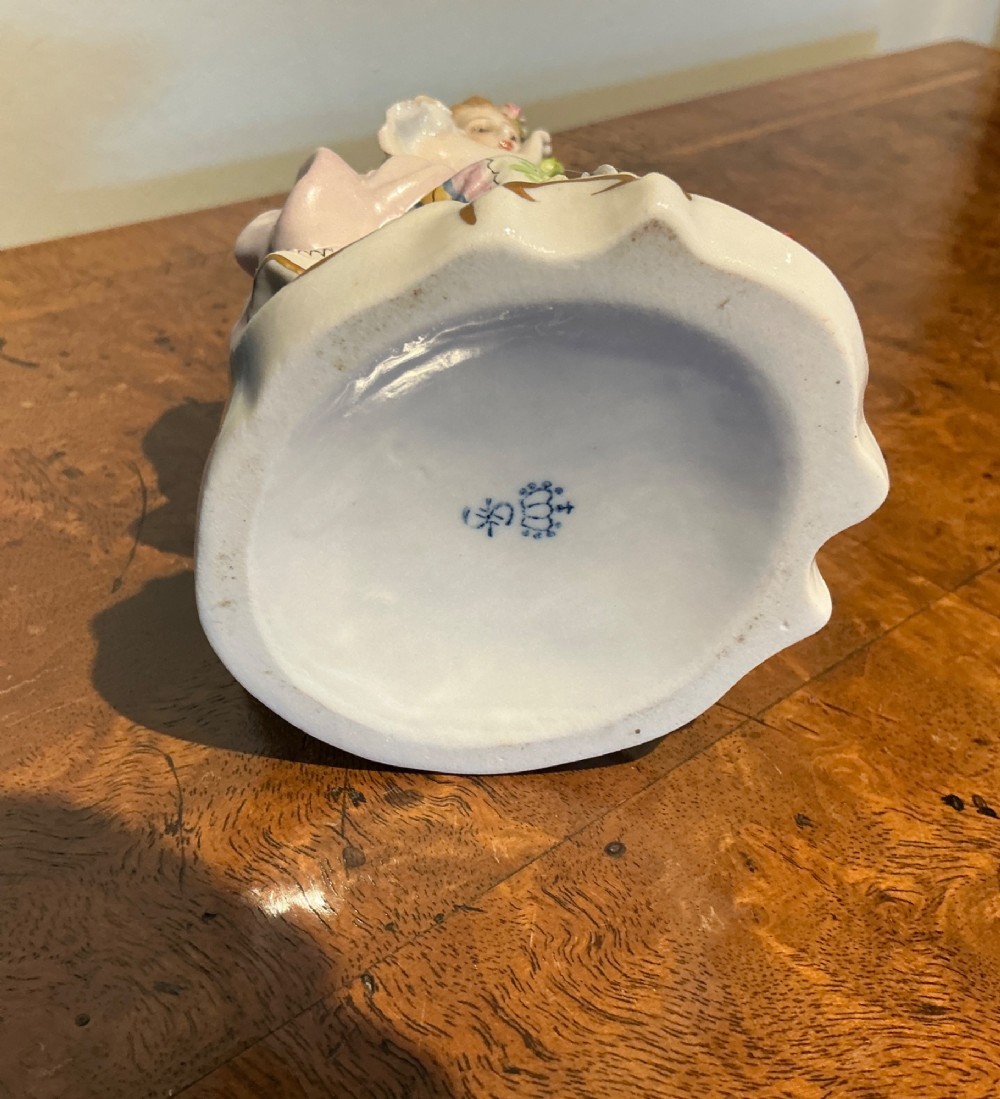 A Sitzendorf porcelain figurine depicting a seated woman in a floral skirt holding a flower-adorned bonnet on a scrolled base. View V