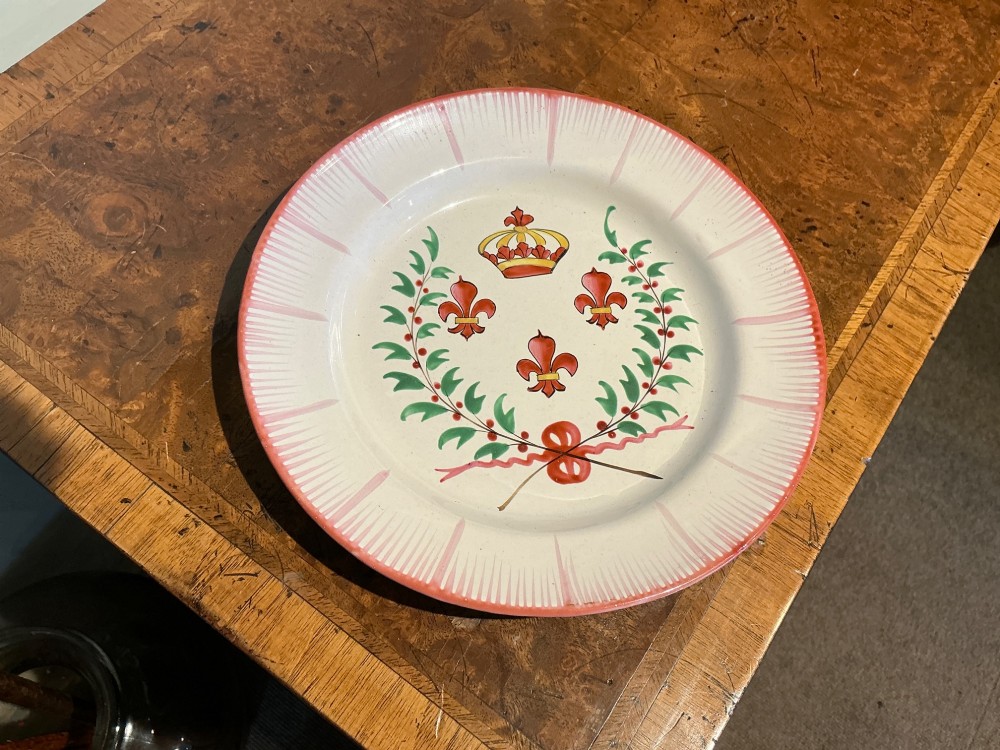 A circular off-white French faience plate decorated with a hand-painted crown, red fleur-de-lis emblems, a green laurel wreath, and a pink patterned rim.
