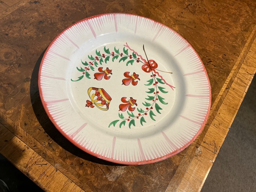 A circular off-white French faience plate decorated with a hand-painted crown, red fleur-de-lis emblems, a green laurel wreath, and a pink patterned rim. View II