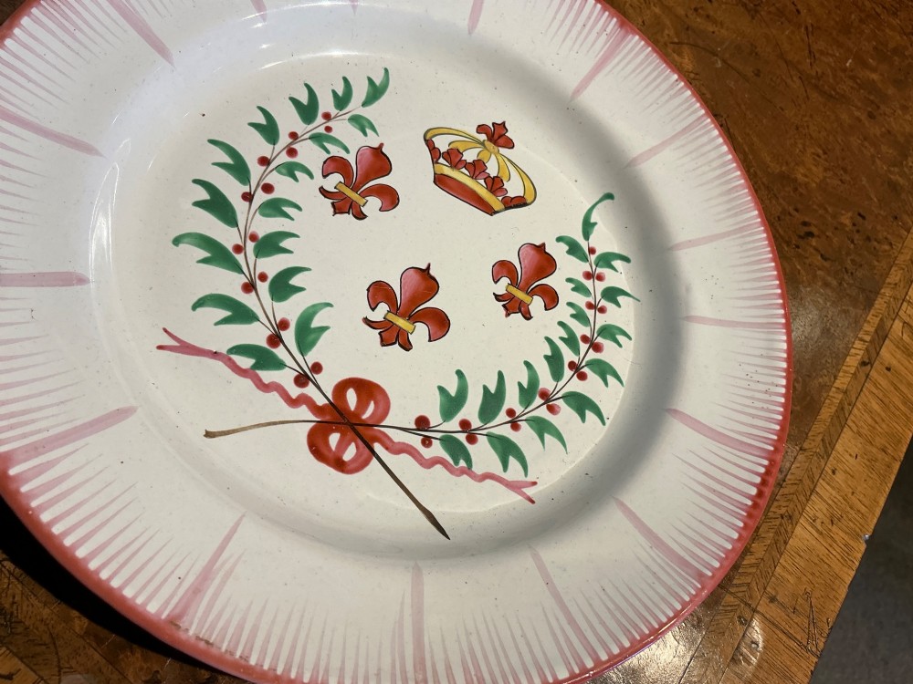 A circular off-white French faience plate decorated with a hand-painted crown, red fleur-de-lis emblems, a green laurel wreath, and a pink patterned rim. View III
