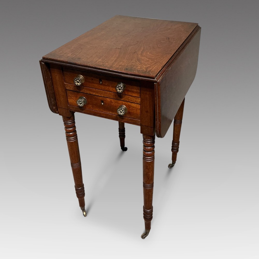 A dark mahogany Georgian Pembroke table featuring two drop leaves, twin drawers with brass knobs, turned legs, and brass castors.