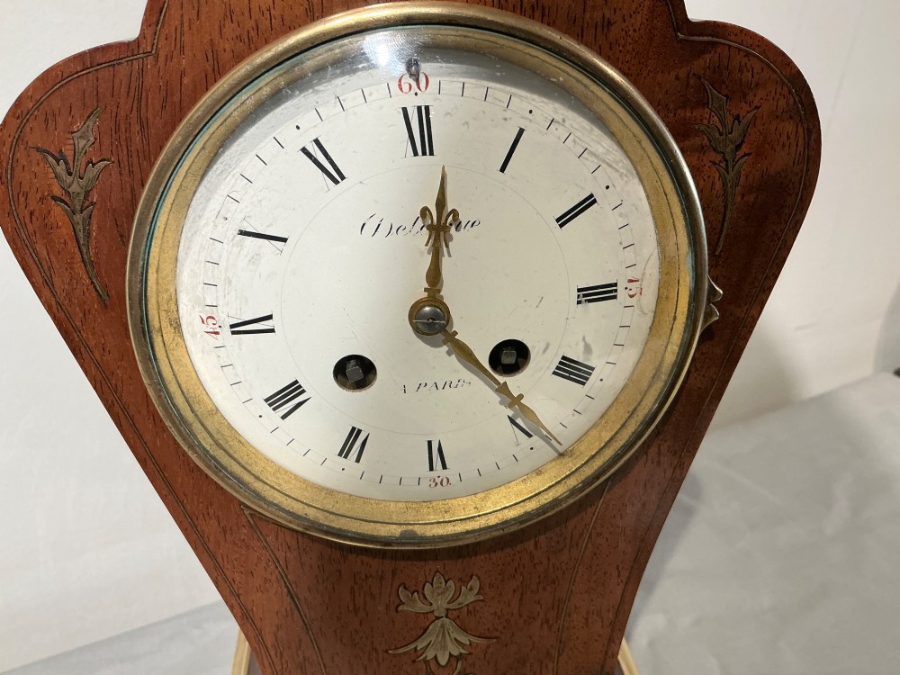 An antique balloon-shaped mahogany mantel clock featuring inlaid brass floral details, a white enamel dial with Roman numerals, and a brass-trimmed plinth base. View VII
