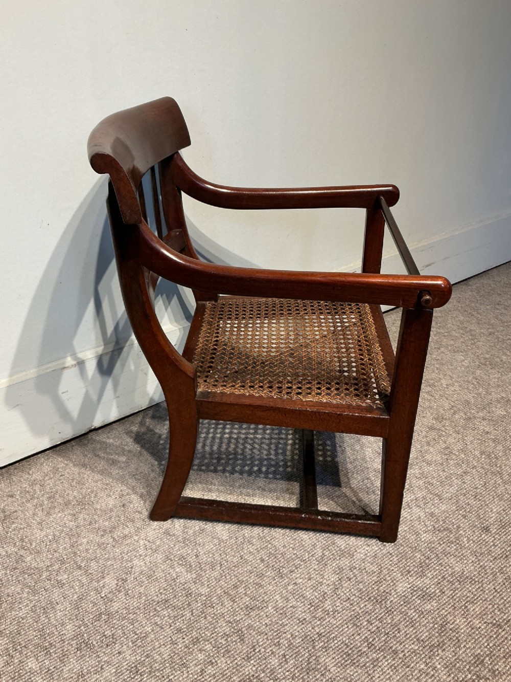 An antique dark mahogany childs armchair featuring a curved top rail, vertical back spindles, a woven cane seat, and a horizontal wooden safety bar. View II