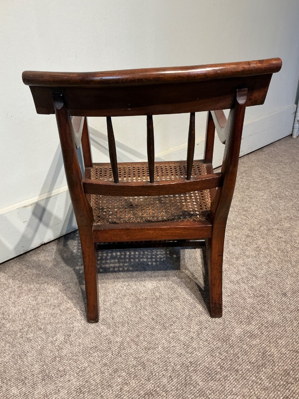 An antique dark mahogany childs armchair featuring a curved top rail, vertical back spindles, a woven cane seat, and a horizontal wooden safety bar. View III