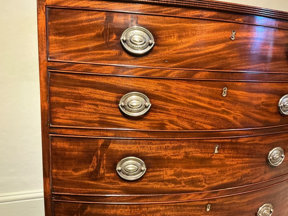 A Georgian flame mahogany bowfront chest of drawers featuring four graduated drawers with oval brass handles and splayed bracket feet. View V