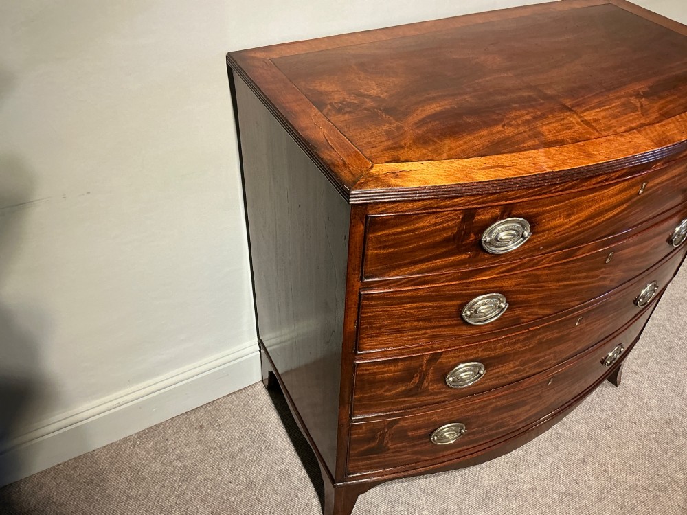 A Georgian flame mahogany bowfront chest of drawers featuring four graduated drawers with oval brass handles and splayed bracket feet. View VI