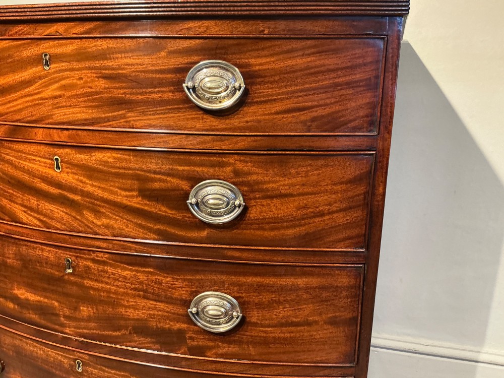 A Georgian flame mahogany bowfront chest of drawers featuring four graduated drawers with oval brass handles and splayed bracket feet. View VII