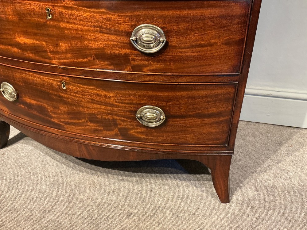 A Georgian flame mahogany bowfront chest of drawers featuring four graduated drawers with oval brass handles and splayed bracket feet. View XII