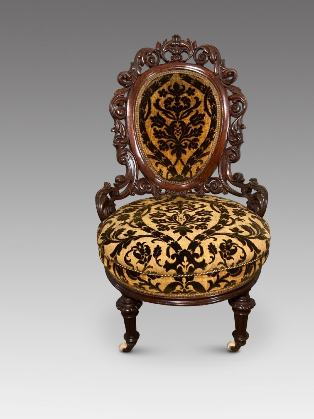 A Victorian mahogany salon chair featuring an ornate carved scrollwork frame, oval backrest, circular seat upholstered in gold and black velvet damask fabric, and turned legs with castors.