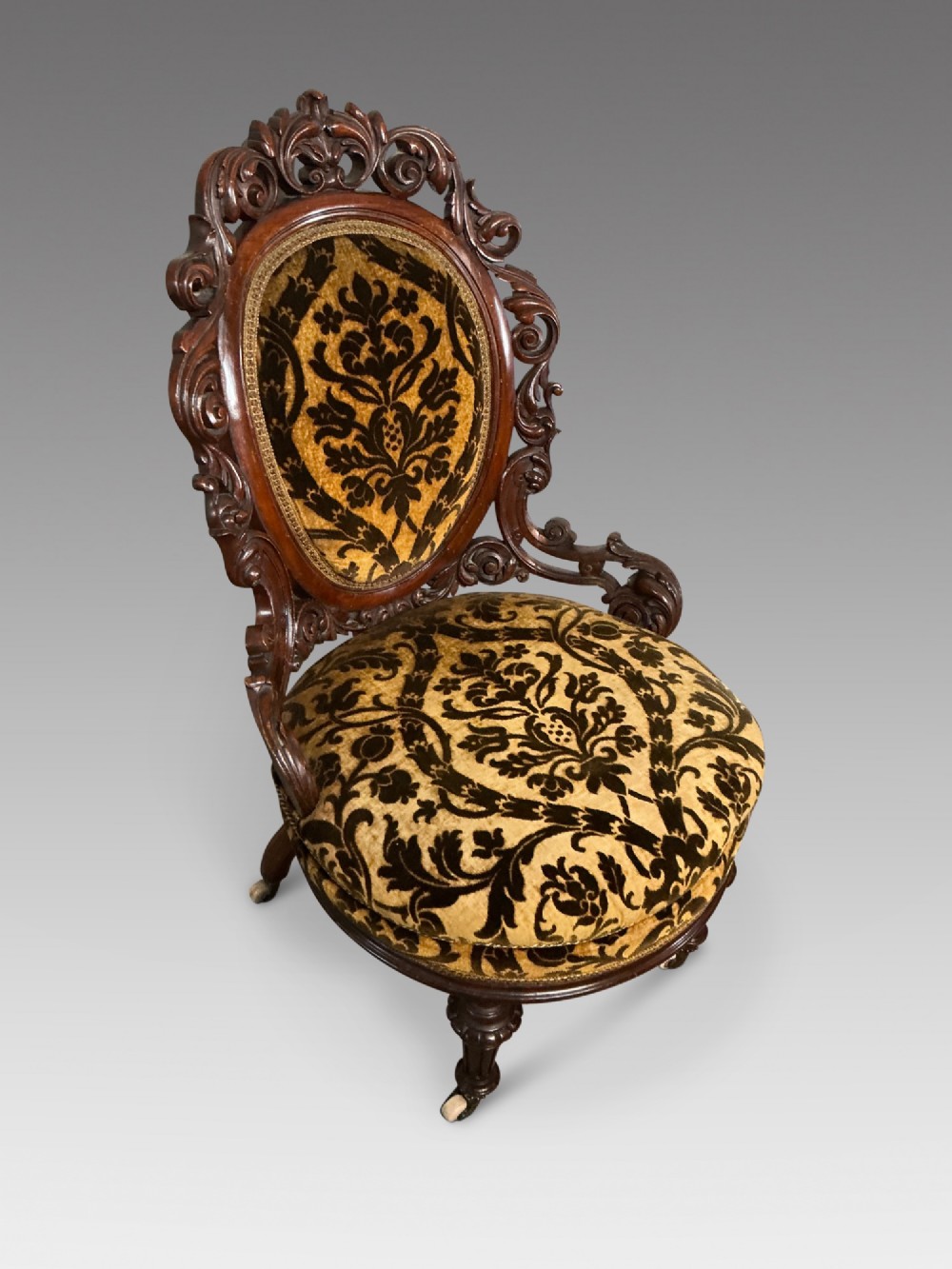 A Victorian mahogany salon chair featuring an ornate carved scrollwork frame, oval backrest, circular seat upholstered in gold and black velvet damask fabric, and turned legs with castors. View II