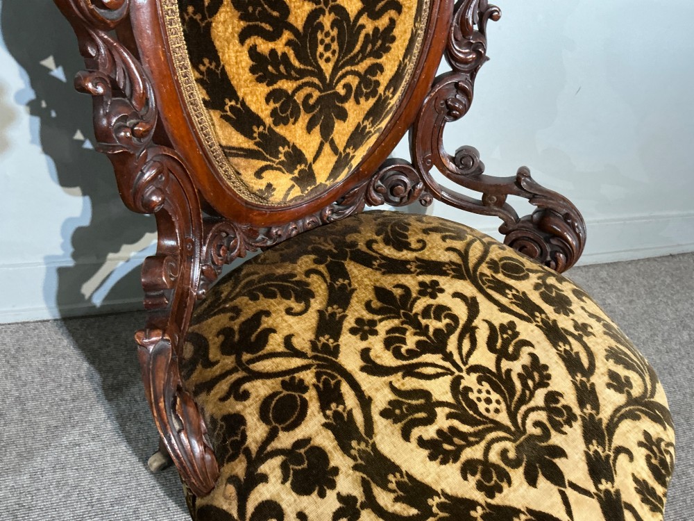 A Victorian mahogany salon chair featuring an ornate carved scrollwork frame, oval backrest, circular seat upholstered in gold and black velvet damask fabric, and turned legs with castors. View V
