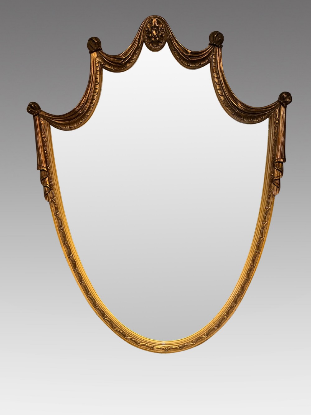 An Edwardian shield-shaped wall mirror featuring a giltwood frame decorated with carved draped swags and a central rosette crest.