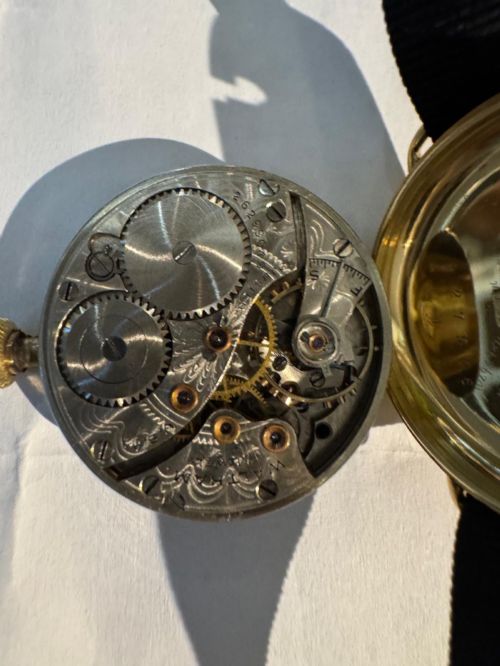 fig 3. A 1920s 18ct gold Waltham wristwatch with a white enamel dial, black Arabic numerals, a red 12 marker, and a black woven strap.