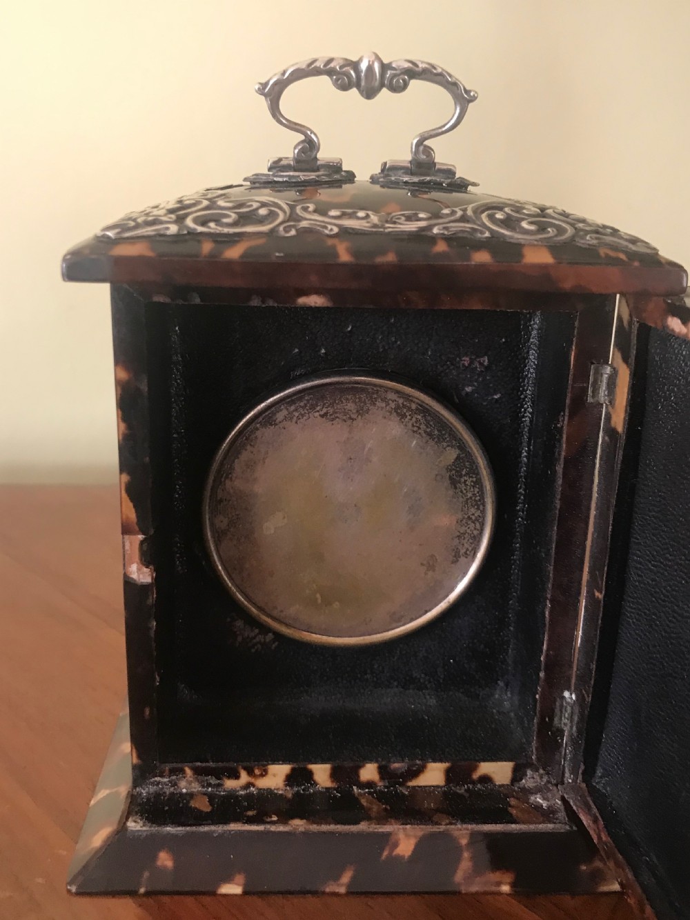 A 19th-century Victorian mantel clock featuring a dark tortoiseshell case, a silver front panel with floral scrollwork, a top carrying handle, and a white circular dial. View V