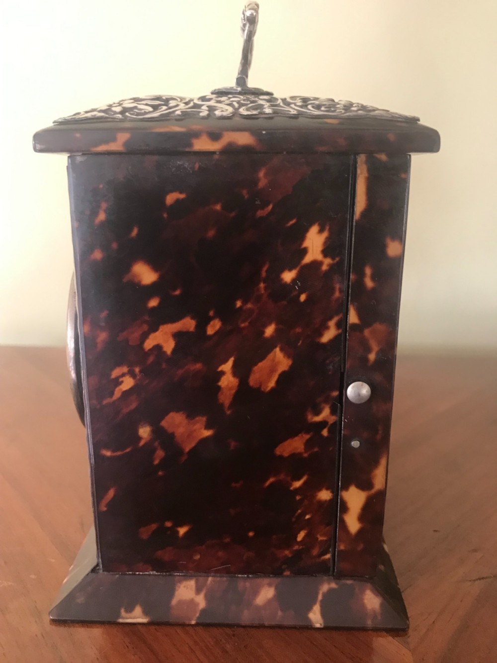 A 19th-century Victorian mantel clock featuring a dark tortoiseshell case, a silver front panel with floral scrollwork, a top carrying handle, and a white circular dial. View VI