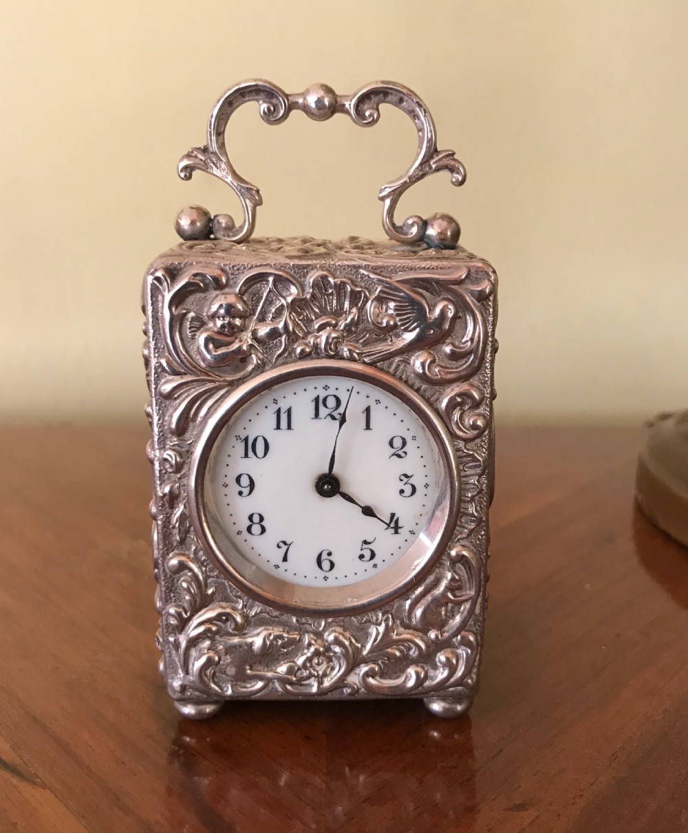A 19th-century miniature silver clock with detailed relief ornamentation, displayed inside an open brown leather travel case. View V