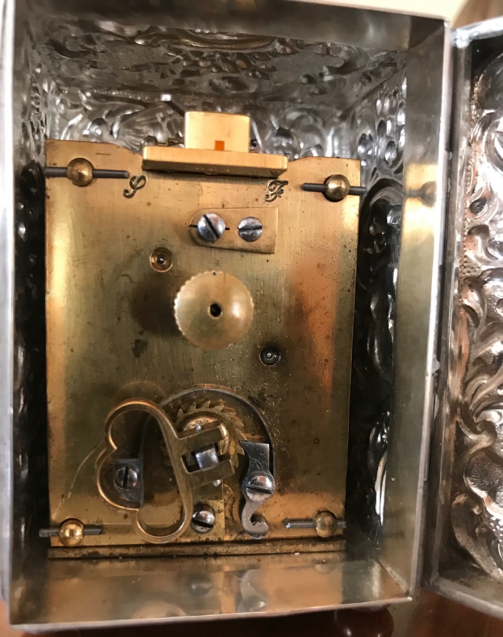 A 19th-century miniature silver clock with detailed relief ornamentation, displayed inside an open brown leather travel case. View VI
