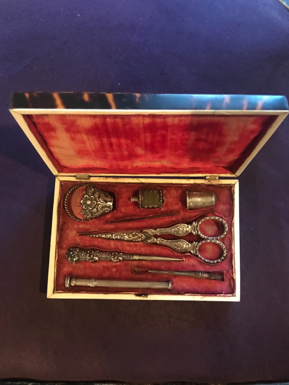 An open Victorian tortoiseshell rectangular sewing box featuring a crimson red velvet fitted interior with intricate silver tools including scissors and a thimble.